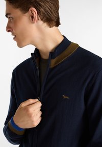 Suéter de cremallera azul marino con cuello acanalado en marrón y azul. Presenta un pequeño logo dorado en el pecho. Textura suave y diseño ajustado.