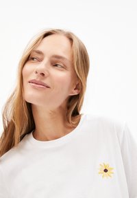Witte katoenen T-shirt met een ronde hals; heeft een gele zonnebloem borduursel op de linkerkant van de borst. Zachte textuur.