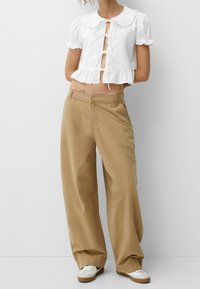 Vit kort blus med volangkrage och puffärmar, ihop med bruna vida byxor. Outfitet inkluderar vita sneakers med svarta accenter.