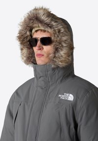 Grå isolerad jacka med en huva klädd i fuskpäls, hög krage och dragkedjefickor. Logotypen är broderad i vitt på vänster bröst.