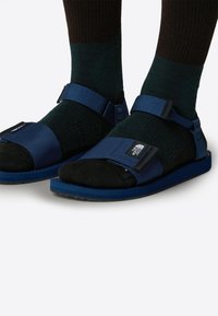 Niebieskie sandały z regulowanymi paskami, czarną amortyzacją i teksturowaną podeszwą. Noszone z ciemnozielonymi skarpetkami w prążki.