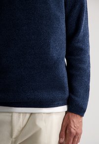 Granatowy sweter o fakturowanej powierzchni z okrągłym dekoltem, z białym wykończeniem i długimi rękawami. Wykonany z dzianiny, prezentujący subtelny wzór.