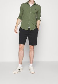 Mężczyzna w oliwkowej koszuli na guziki, czarnych szortach, białych skarpetkach i białych sneakersach stoi na tle jasnego, jednolitego tła.