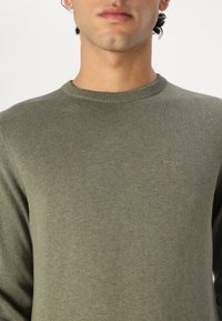 Olíva zöld kötött pulóver kerek nyakkal és finom logóval a mellrészen. Simaság jellemzi a textúrát és a testhez simuló fazont, kiemelve a stílus egyszerűségét.