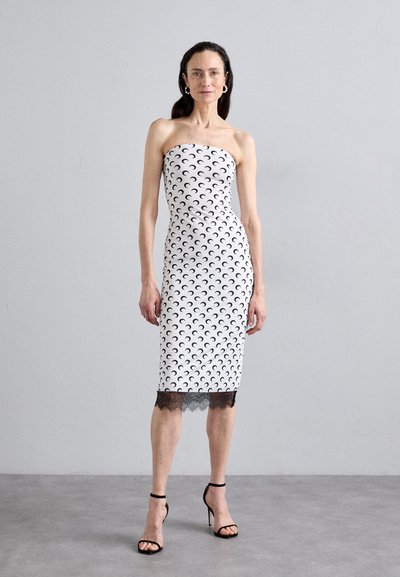 Vestido blanco sin tirantes con patrones de media luna negra, de silueta ajustada, a la altura de la rodilla, terminado con un dobladillo de encaje negro, combinado con sandalias de tacón negro.