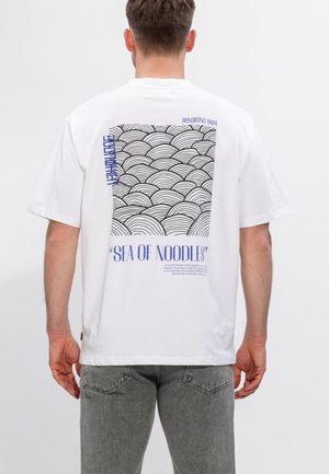 Mand iført hvid T-shirt med sort bølgemønster og blå tekst "SEA OF NOODLES" på ryggen, kombineret med grå jeans.