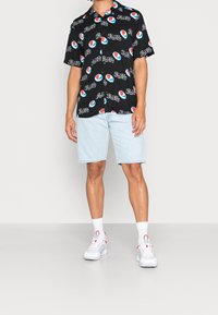 Black short-sleeve shirt with repeated Pepsi logo pattern, paired with light blue denim shorts and white sneakers with red accents.