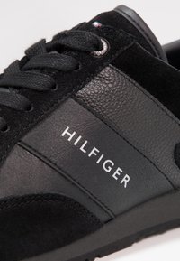 Scarpa da ginnastica nera con pannelli in pelle testo e liscia, dettagli in suede, lacci neri e la scritta "HILFIGER" stampata sul lato.