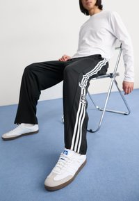 Melni sporta bikses ar baltām svītrām, ar logotipa detaļu, apvienotas ar baltām kedām ar ziliem akcentiem. Modelis sēž uz hroma krēsla.