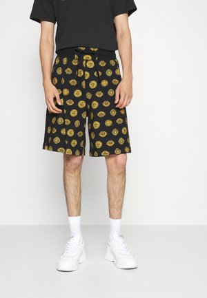 Homme portant un short noir avec des motifs circulaires jaunes, un t-shirt noir, des chaussettes blanches et des baskets blanches, debout devant un fond uni.