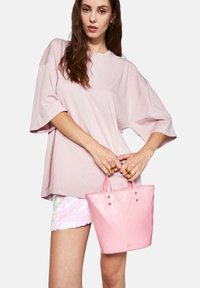 Rosa tygväska med geometriskt mönster, glänsande yta, två små handtag och guldfärgade detaljer. Bärs med en lös rosa skjorta och paljettshorts.