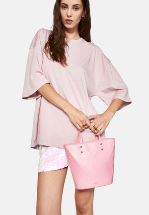 Lyserød totebag med et geometrisk mønster, glat tekstur, to små håndtag og gyldne detaljer. Båret med en løs lyserød skjorte og palietshorts.