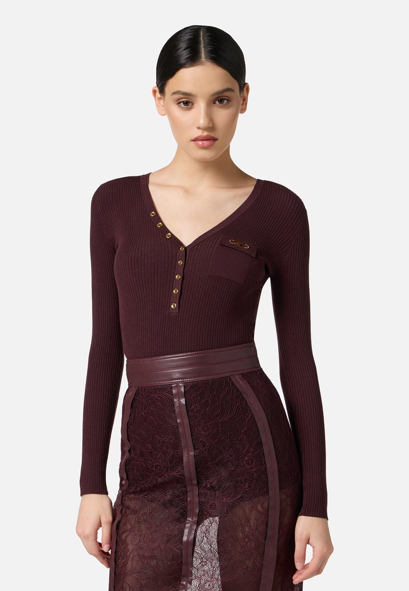 Top lungo a maniche con costine color bordeaux, scollo a V, caratterizzato da bottoni decorativi in oro e una piccola tasca. Abbinato a una gonna in pizzo testurizzato.