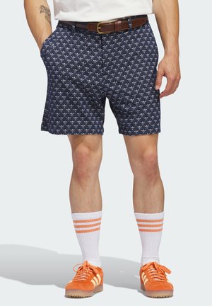Mann trägt marineblaue gemusterte Shorts mit braunem Gürtel, weiße Socken mit orangen Streifen und orangefarbene Sneakers mit weißen Streifen.