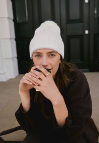 Bela pletena kapa s cofom, temen plašč, drži enkratno kavna skodelico. V ozadju je trdna temna vrata.