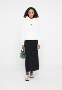 Haut à capuche blanc avec logo, jupe maxi noire à couches avec un design angulaire, baskets grises et un petit sac à main vert.