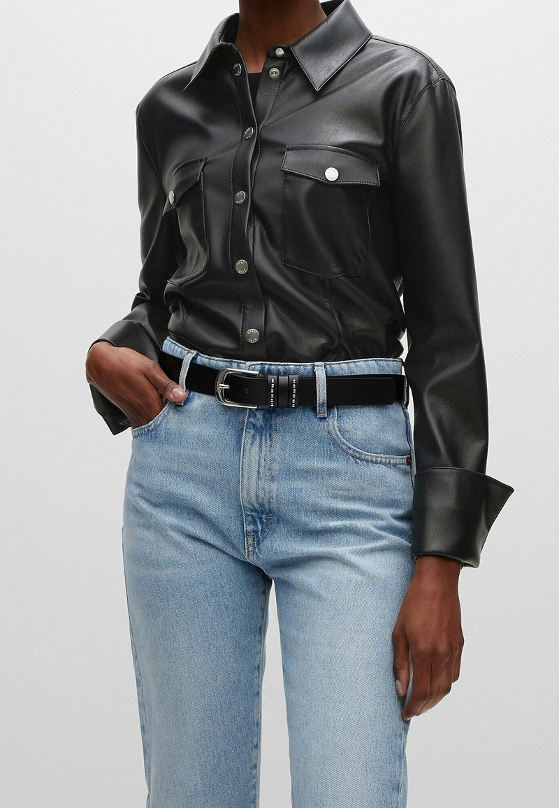 Chemise en cuir noir à boutons avec deux poches avant, associée à un jean en denim bleu clair et une ceinture noire. Texture lisse et coupe ajustée.