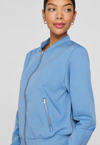 Svetlo modra bomberska jakna iz gladkega blaga, ki ima zlat zadrgo in stranske žepe ter rebrast ovratnik in manšete.