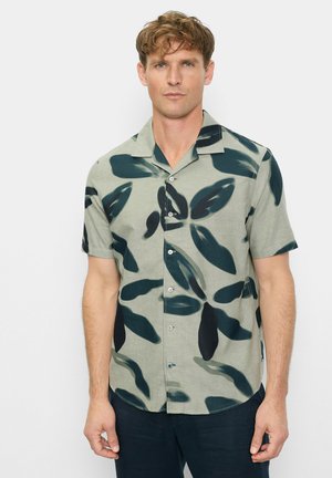 Man met kort golvend haar, gekleed in een kort mouwig overhemd met een grote donkergroene bladprint en een donkere broek, staand tegen een effen achtergrond.