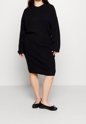 Femme portant une robe-pull noire à manches longues et des ballerines noires, debout devant un fond clair uni.