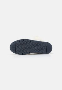 Sål til fodtøj i mørkeblå gummi med et tekstureret grebsmønster. Det lyse beige overmateriale er delvist synligt. Flad, rund form.