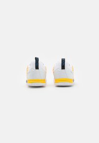 Vista trasera de zapatillas deportivas blancas con acentos naranjas, suelas amarillas, diseño texturizado y tiradores azul oscuro en el talón.