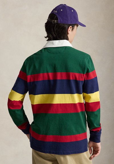 Άνδρας με την πλάτη, φοράει μακρυμάνικο ράγκμπι πουκάμισο με οριζόντιες ρίγες πράσινου, κόκκινου, ναυτικού και κίτρινου χρώματος και μια μοβ μπέιζμπολ καπέλο.