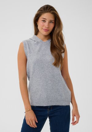 Jeune femme aux longs cheveux bruns portant un haut en maille gris clair sans manches et un jean bleu foncé, debout devant un fond clair uni.