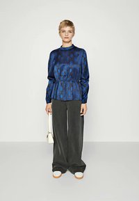 Blusa peplum in seta blu con motivo e maniche lunghe, abbinata a pantaloni grigi a gamba larga e sneakers bianche con accenti arancioni.
