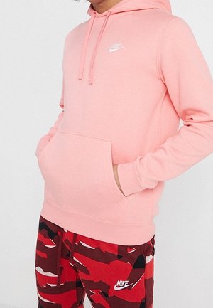 Osoba ubrana w jasnoróżową bluzę z kapturem Nike z dłońmi w przedniej kieszeni oraz czerwono-czarno-białe spodnie moro Nike na jednolitym tle.