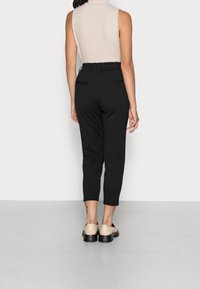 Pantalons noirs coupés avec une taille élastique, deux poches arrière et une texture lisse. Portés avec un haut sans manches beige clair.