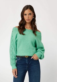 Maglione verde menta off-shoulder con polsini a coste e dettagli in pizzo sulle maniche, abbinato a jeans blu scuro e una cintura marrone con hardware dorato.