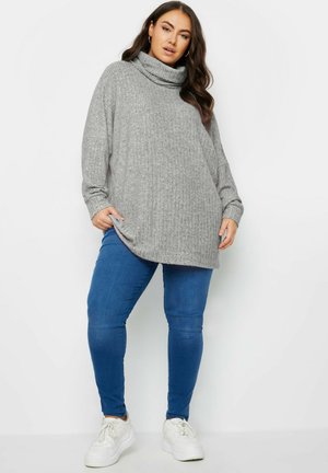 Grauer, gerippter Rollkragenpullover mit langen Ärmeln, kombiniert mit blauen Slim-Fit-Jeans und weißen Sneakers. Der Saum ist an den Seiten asymmetrisch.