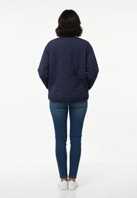 Frau von hinten, die eine marineblaue gesteppte Jacke, enge blaue Jeans und weiße Sneakers auf einem schlichten weißen Hintergrund trägt.