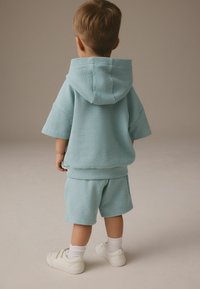Světle modrá texturovaná mikina s kapucí, krátkými rukávy, odpovídající šortky a bílé tenisky, z pohledu zezadu. Jednoduchý design, měkká látka.