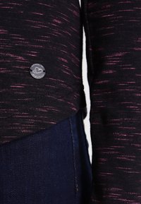 Nahaufnahme von dunkel gemarcktem Stoff mit kleinem rundem schwarzen Logo-Patch, das ein Herz zeigt, getragen über dunkelblauen Jeans.