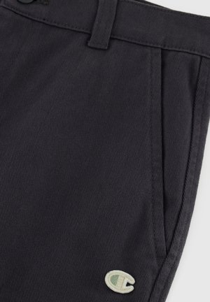 Pantalones de textura gris oscuro con un solo pliegue, trabillas para cinturón y un pequeño logo bordado en verde claro en la parte inferior izquierda.