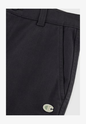 Pantaloni grigi scuri con texture, caratterizzati da una piega singola, passanti per cintura e un piccolo logo ricamato verde chiaro sul lato sinistro inferiore.