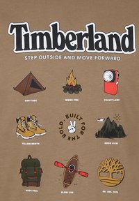 Brązowa koszulka z graficznymi nadrukami: namiot, ognisko, lampka kieszonkowa, żółte buty, plecak, kajak, góry, logo drzewa oraz hasło.