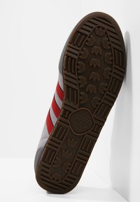 Grå sneakers med röda ränder, gummisula, strukturerad yta och distinkt logotyp på sulan. Formade för komfort och grepp.