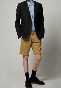 Musta bleiseri vaaleansinisen nappipaidan päällä, yhdistetty khakin värisiin shortseihin. Asun täydentävät mustat nahkakengät ja mustat pilkulliset sukat.