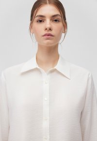 White button-up shirt with a classic collar. Made of lightweight, textured fabric. Features pearl-like buttons along the front placket.