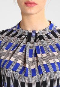 Närbild av en person som bär en blus med hög hals och ett geometriskt mönster i blått, svart, beige och vitt, där endast läppar och nacke syns.