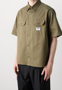 Olivgrünes kurzärmeliges Hemd mit durchgehender Knopfleiste, zwei Brusttaschen, silbernen Knopfdetails und glatter Stoffstruktur. Weißes Etikett angebracht.
