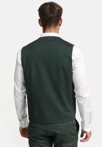 Grønn fiskebeinsvest laget av strukturert stoff, med en hvit skjorte med krage under. Har en tettsittende design og ingen synlig maskinvare.