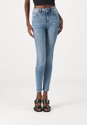 Slim fit -malliset haalistuneet siniset farkut, joita on yhdistetty mustiin avoimilla varpailla varustettuihin sandaaleihin, joissa on kullanväriset solkikoristeet, kuvattuna vyötäröstä varpaisiin.