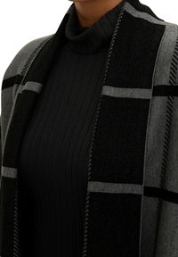 Schwarz-grau karierter Schal mit weicher Textur und fransigen Kanten, über einem gerippten schwarzen Rollkragenpullover geschichtet.