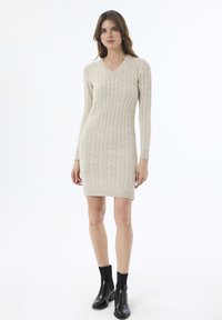 Beige gebreide jurk met een V-hals, lange mouwen en een textuur van kabelpatroon. Gepaard met zwarte enkellaarsjes en een effen achtergrond.