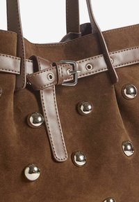 Barna velúr bevásárlótáska ezüst színű szegecsekkel és sima bőrszíjjal, csattal díszítve, kiemelve a rendezett formáját.