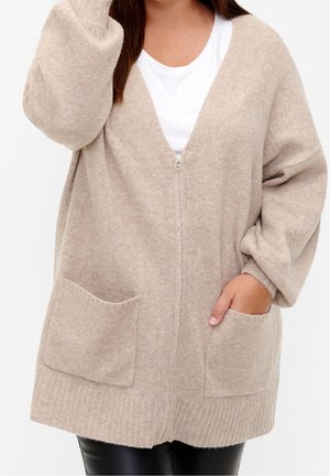 Beige gebreide cardigan met een V-hals, frontale rits en twee grote zakken. Heeft brede mouwen en een geribbelde zoom en manchetten. Soepele textuur.